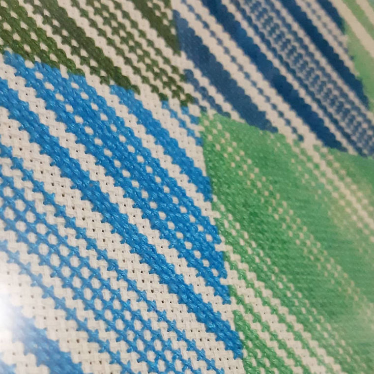 Closeup of a cross stitch piece of grid squares in blue and green colours. The squares comprise many diagonal lines in various widths, all running from the top left of the frame to the bottom right.