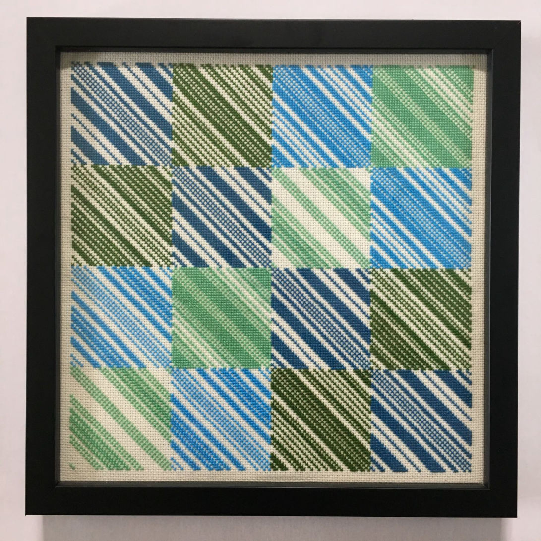A framed cross stitch piece of a four-by-four square grid in alternating colours of blue and green. Each grid square comprises many diagonal lines in various widths, all running from the top left to bottom right.