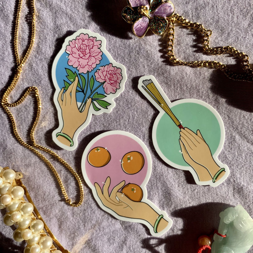Three stickers grouped together on a pale purple surface surrounded by pieces of jewellery. The stickers all show a beige hand wearing a smooth green bracelet in various poses: reaching towards two pink flowers, juggling three tangerines, and pressed together in prayer, holding a bundle of incense sticks.