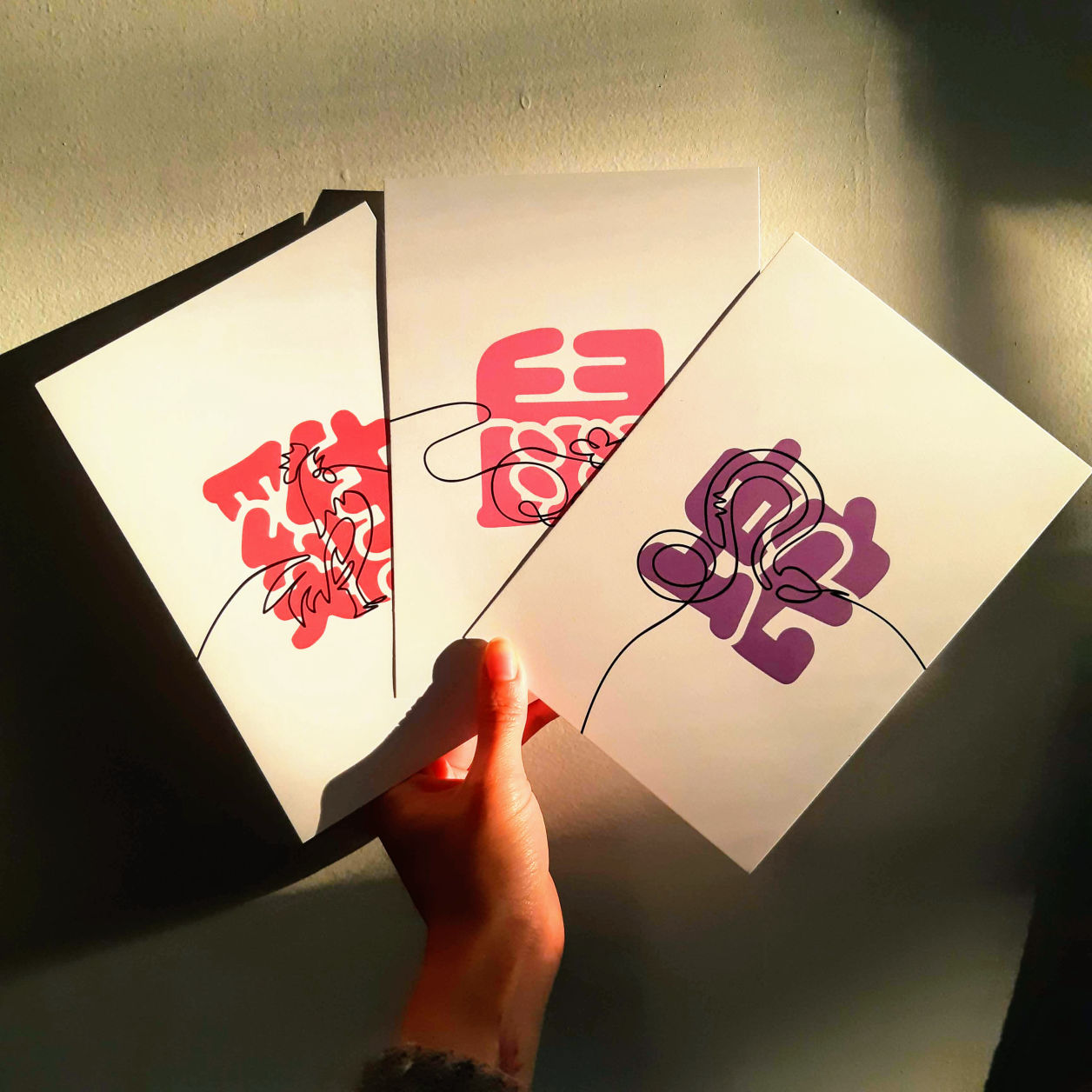 Three postcard art prints held up to the sunlight by a light beige hand. Each card features a line drawing of an animal and a bubble lettered Chinese character, from left to right: Rooster, Rat, and Snake.